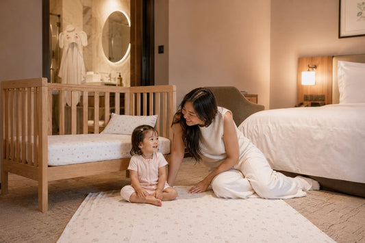 Mother and toddler playing on a soft bamboo blanket beside a wooden baby cot with bamboo sheets in a family-friendly hotel room.